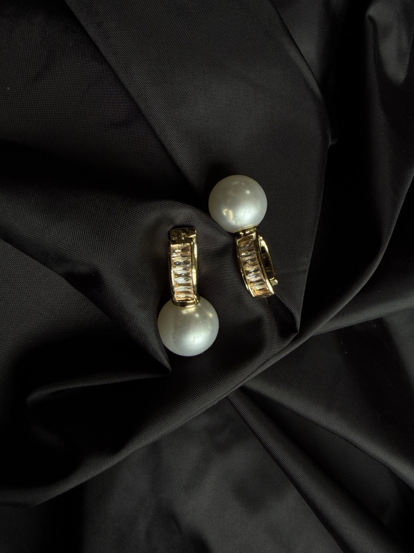A pair of gold hoop earrings with large white pearl drops, displayed on a draped black satin fabric background.
