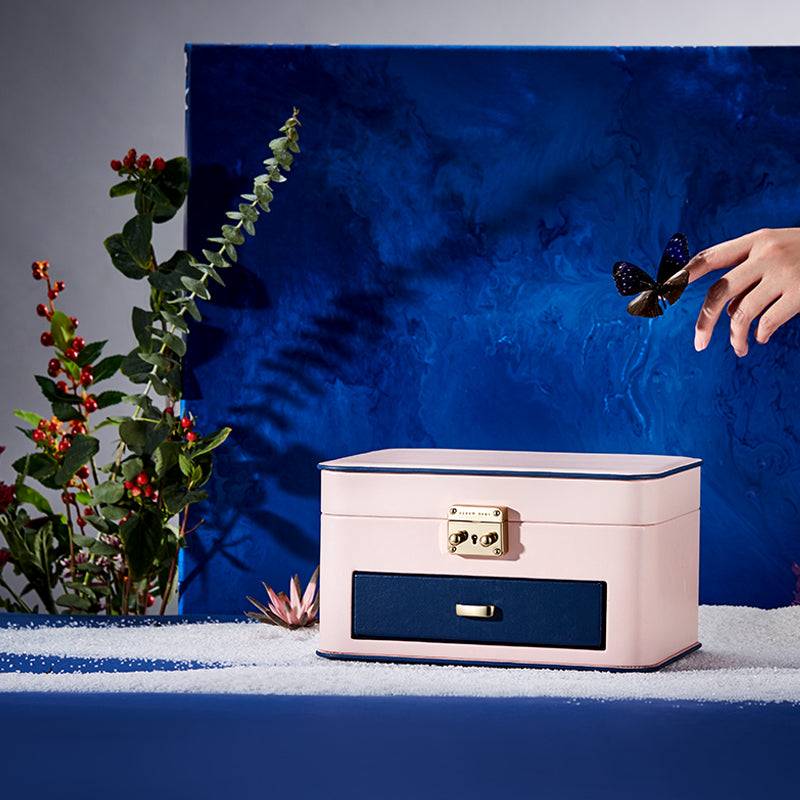 Jewelry storage box in pink with navy blue drawer and gold latch on a blue backdrop.