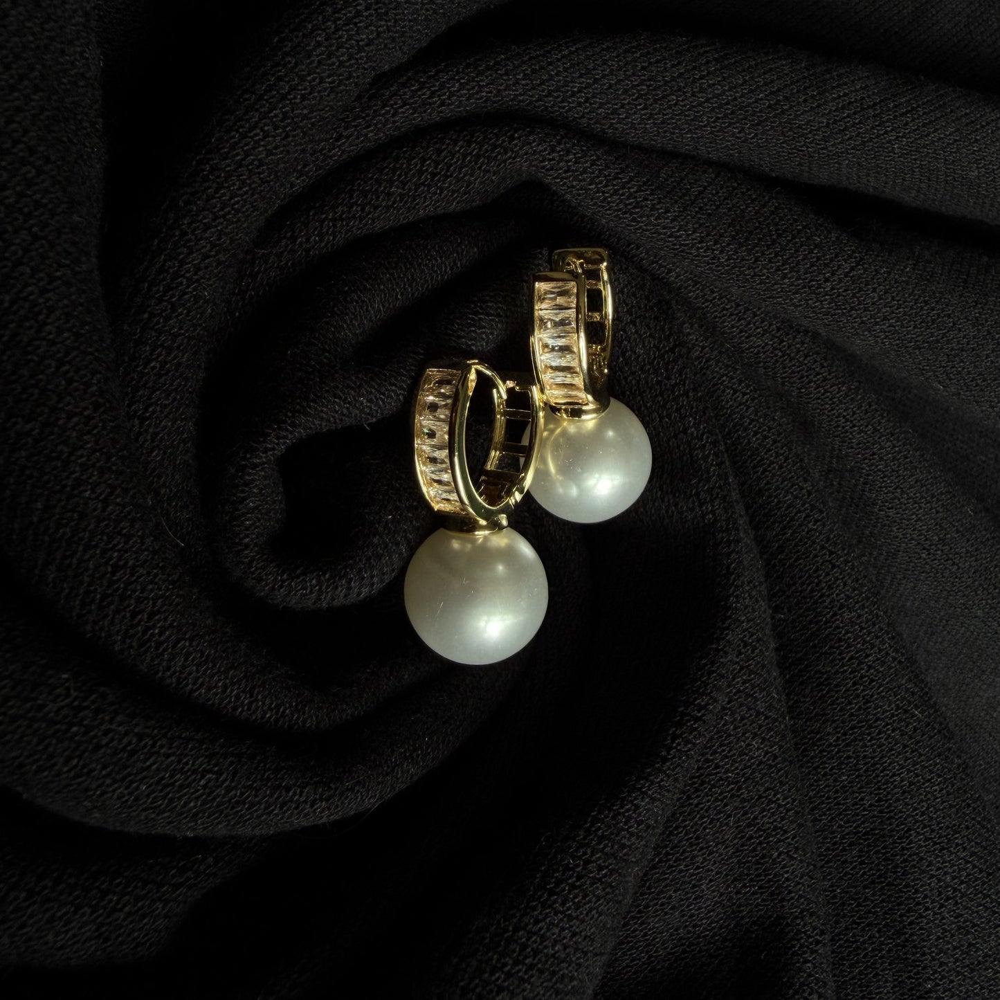 A pair of gold hoop earrings with white pearl drops, displayed on a textured black fabric background.