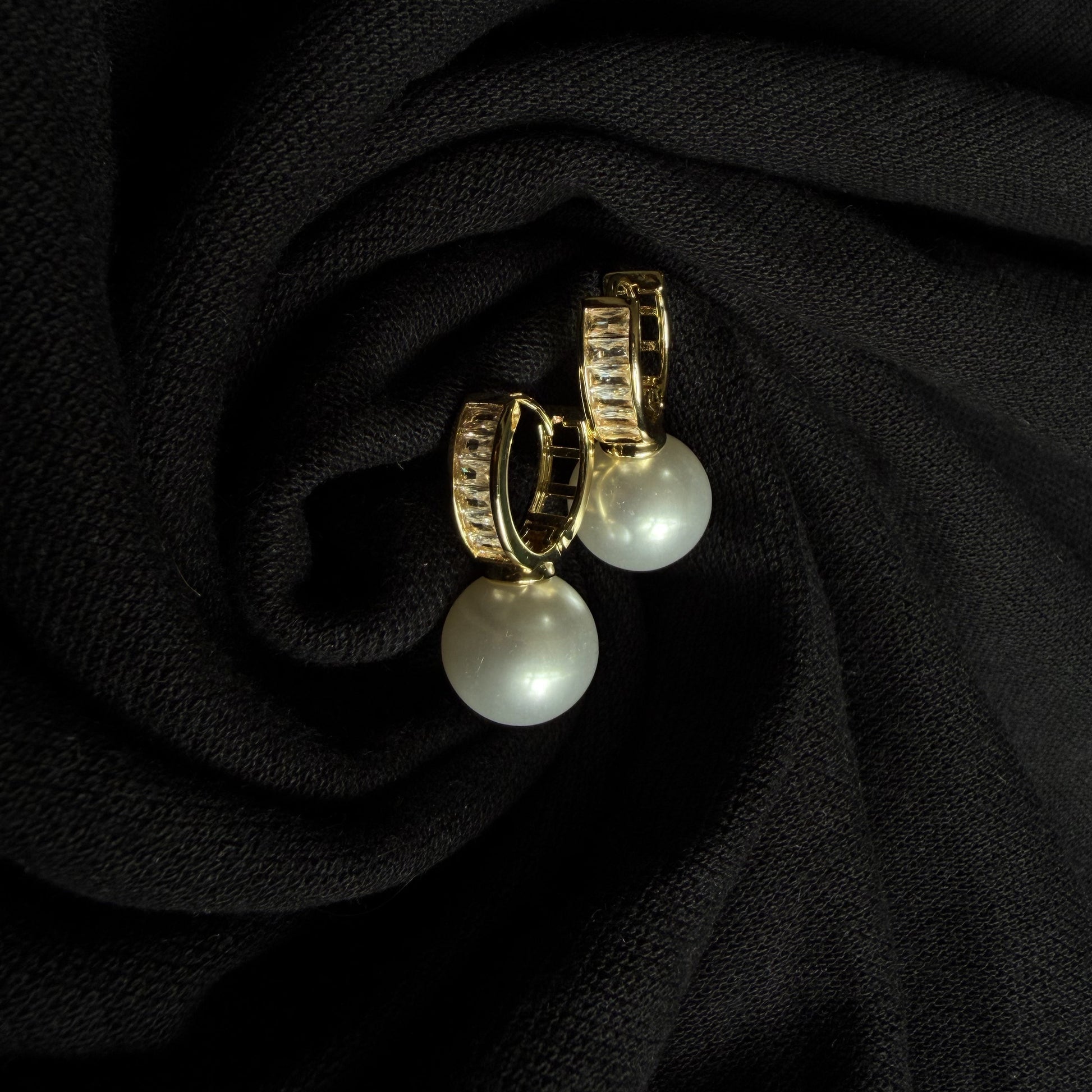 A pair of gold hoop earrings with white pearl drops, displayed on a textured black fabric background.