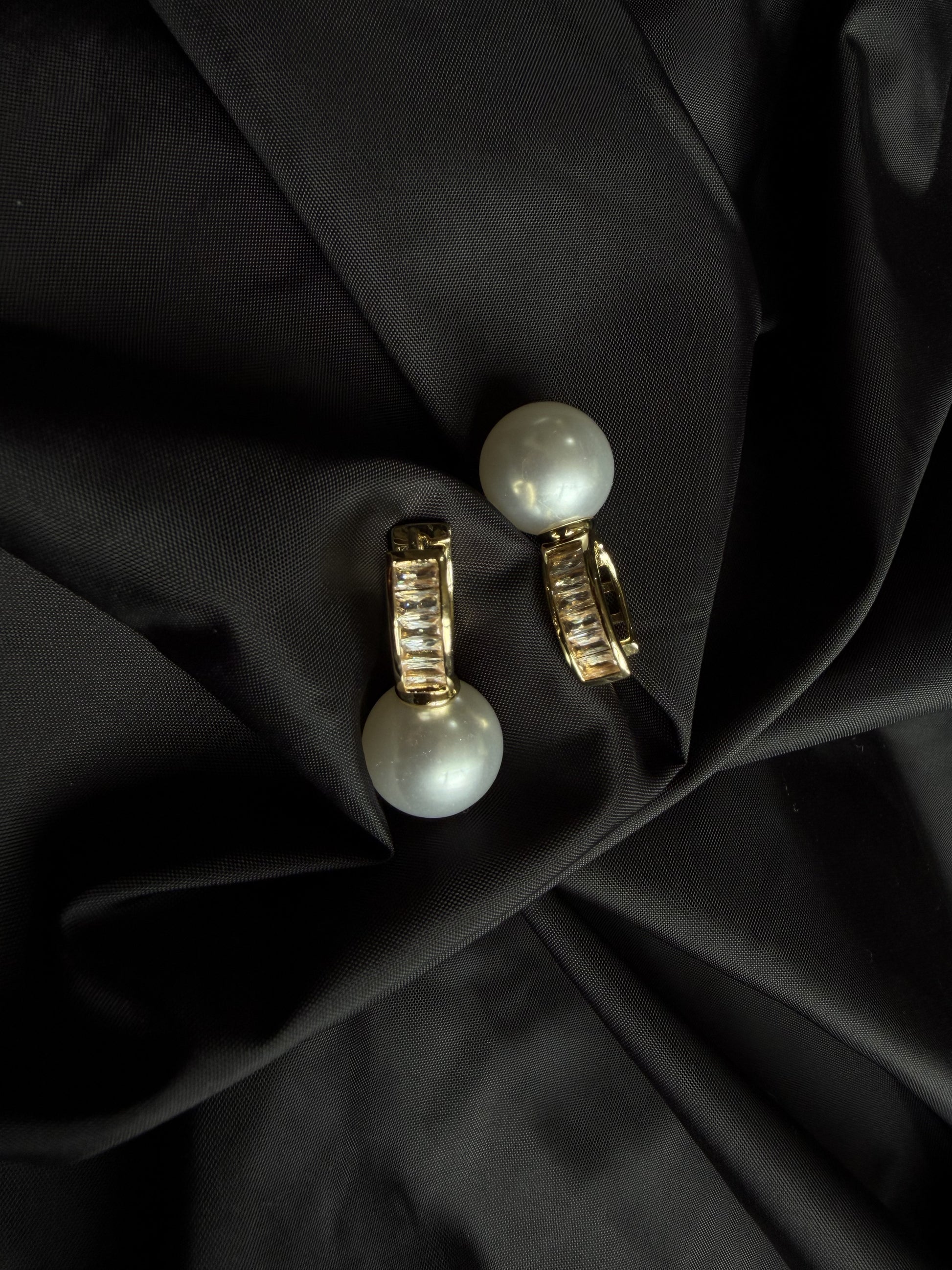 A pair of gold hoop earrings with large white pearl drops, displayed on a draped black satin fabric background.