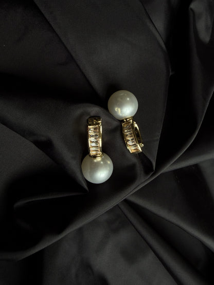 A pair of gold hoop earrings with large white pearl drops, displayed on a draped black satin fabric background.