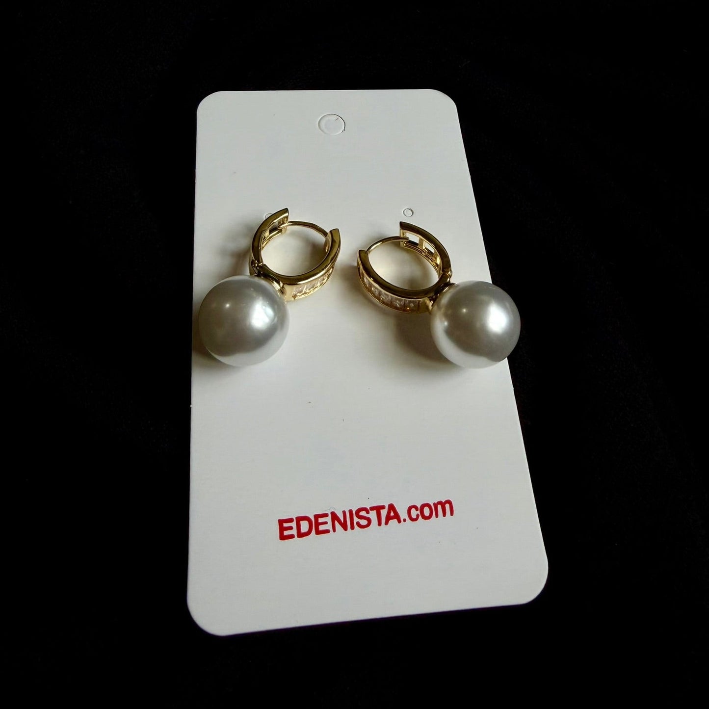 A pair of gold huggie hoop earrings with white pearl drops, displayed on a white card labeled 'EDENISTA.com' against a black background.