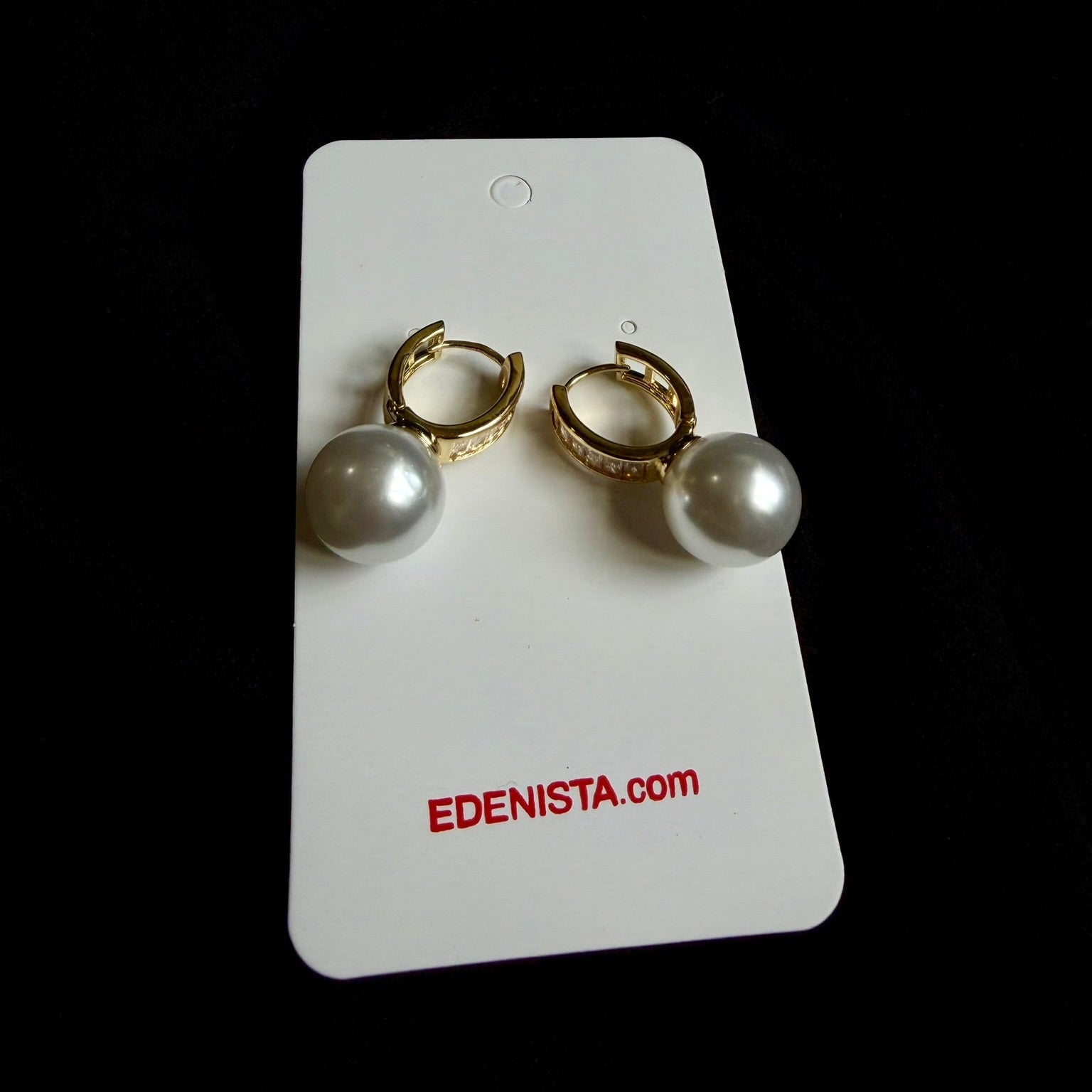A pair of gold huggie hoop earrings with white pearl drops, displayed on a white card labeled 'EDENISTA.com' against a black background.