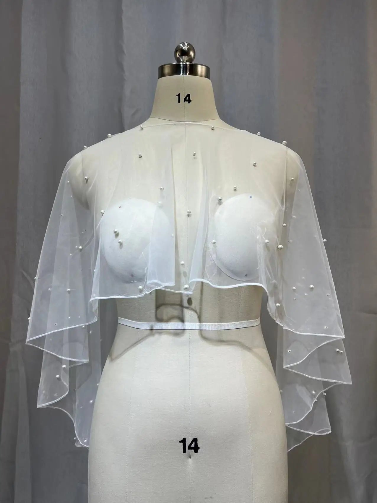 Dressmaker mannequin displaying sheer white tulle pearl-embellished bridal cape bolero with two-layer flared hem