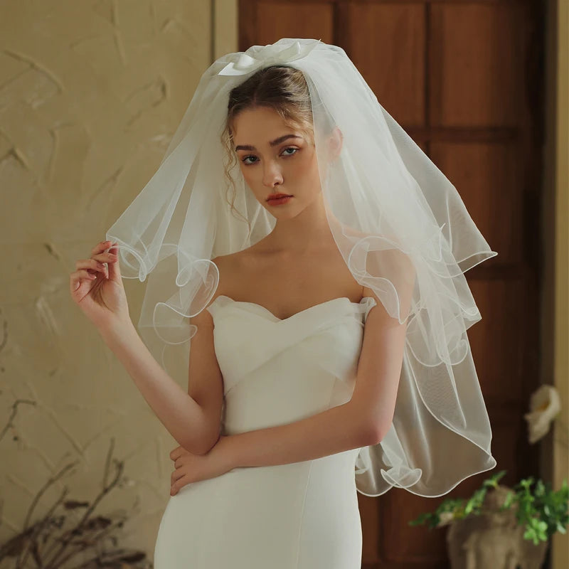 Model holding edge of short two-tier white bridal veil with curled ruffled layers and satin bow hair clip