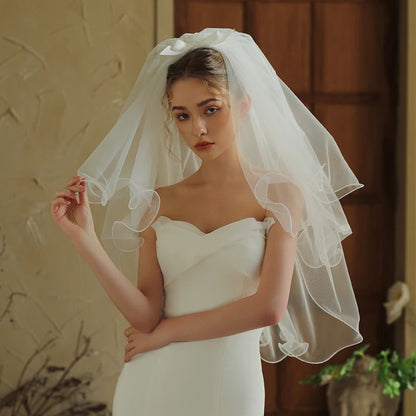 Model holding edge of short two-tier white bridal veil with curled ruffled layers and satin bow hair clip