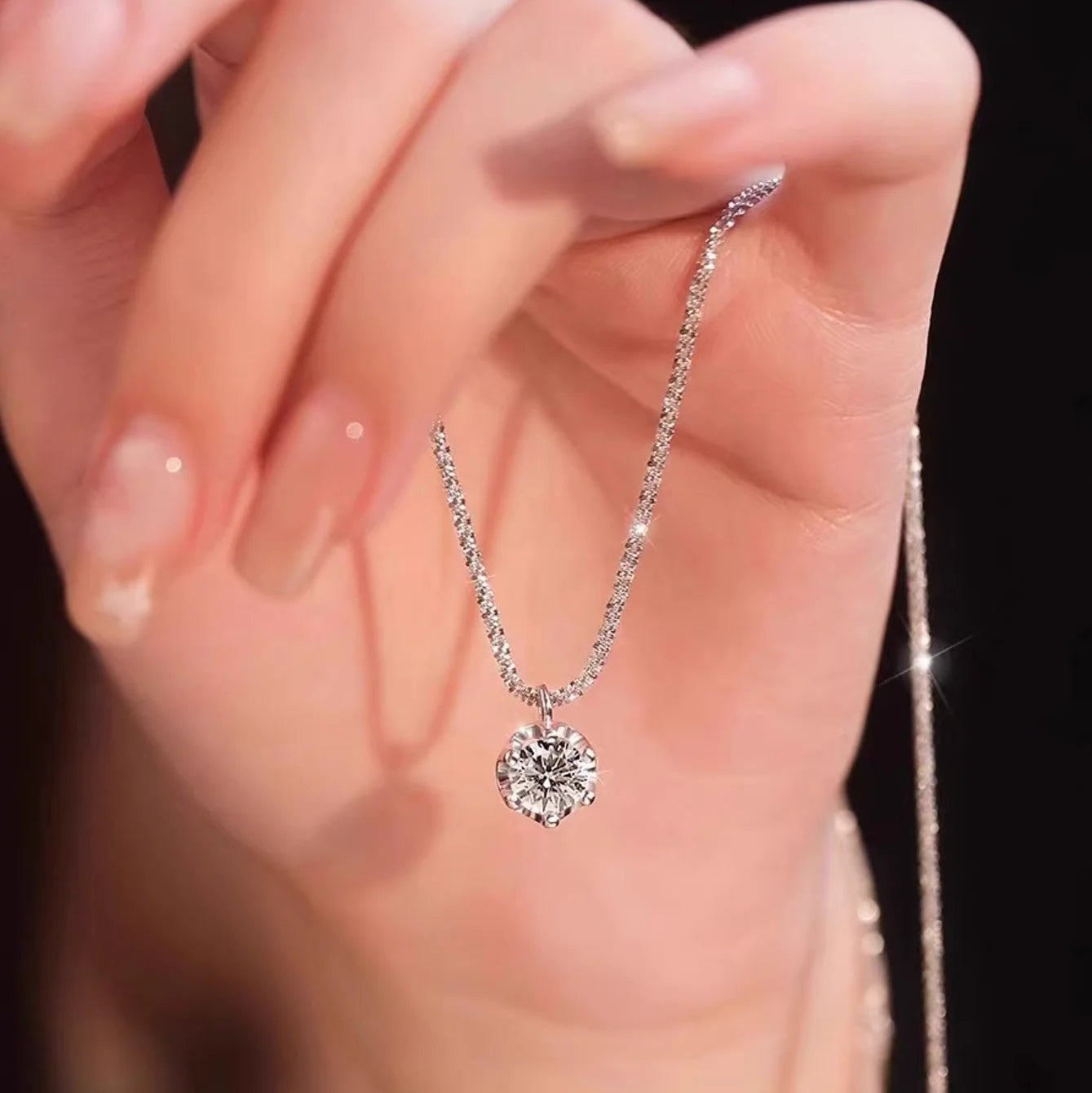 Hand holding sparkling silver moissanite solitaire pendant necklace with glittering chain against dark background