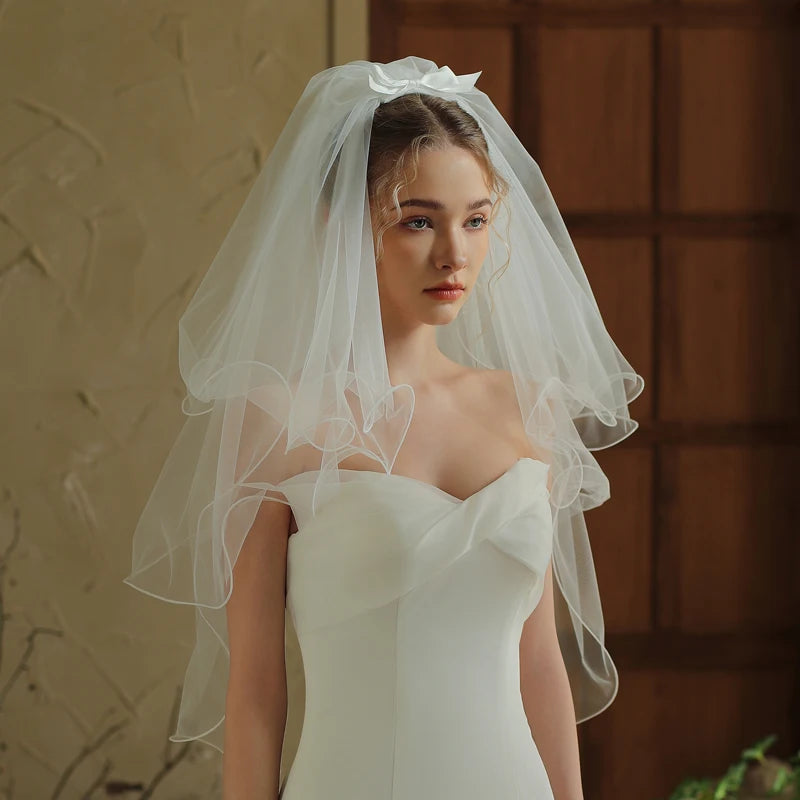 Front view of model wearing short two-tier white bridal veil with curled ribbon edge and satin bow at crown over white strapless gown