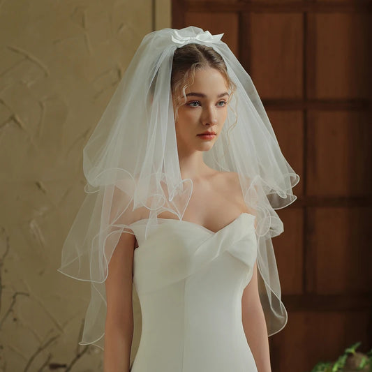 Front view of model wearing short two-tier white bridal veil with curled ribbon edge and satin bow at crown over white strapless gown