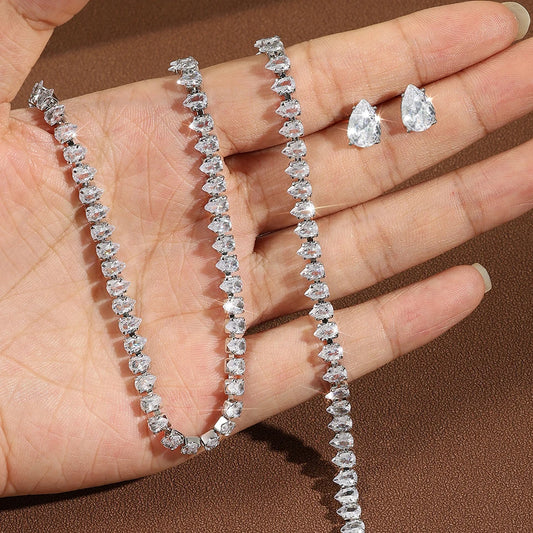 Open palm displaying silver pear-cut CZ water drop tennis necklace and matching bracelet with pear-shaped stud earring on brown background