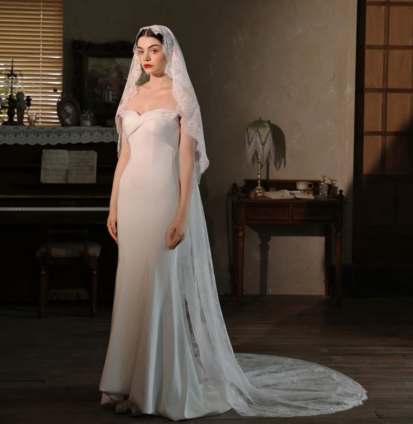 Full-length front view of bride in white strapless wedding gown with long white lace chapel veil in vintage-styled room with piano